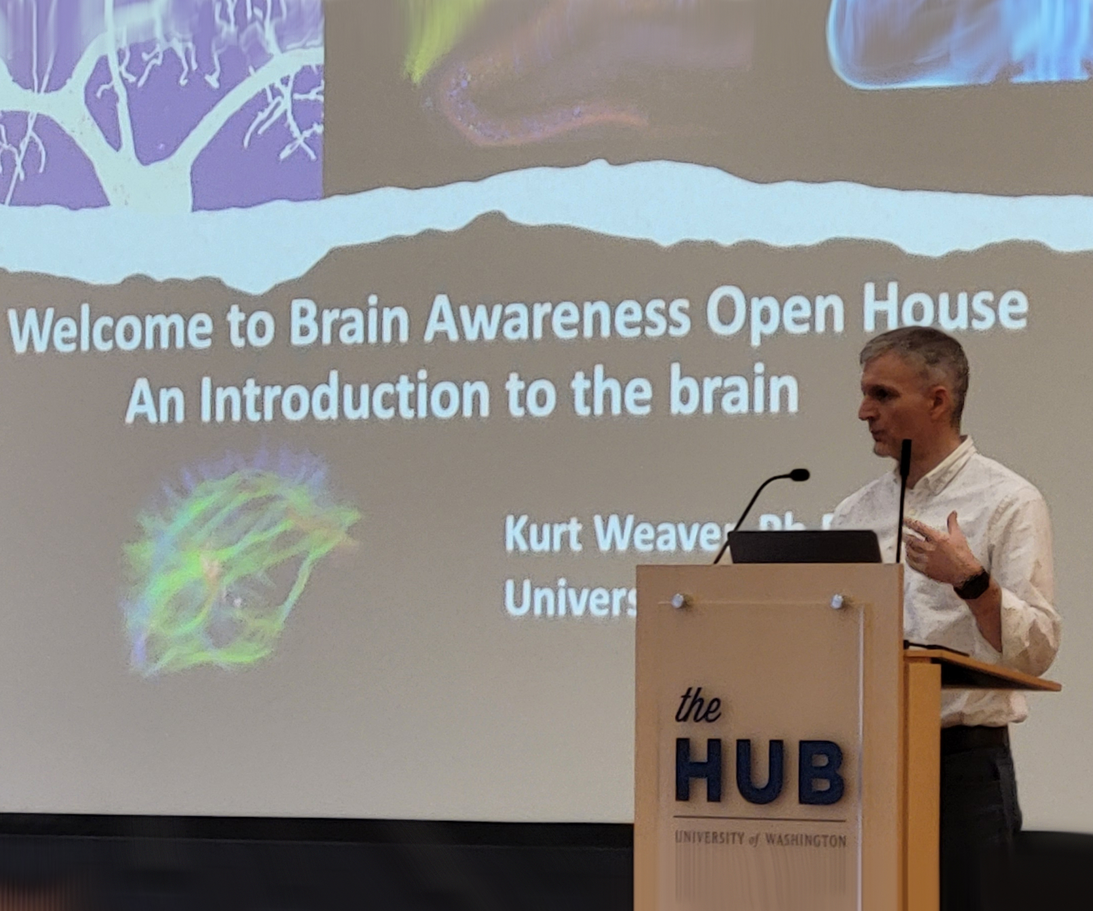 Kurt Weaver standing at a podium in front of a screen reading Welcome to Brain Awareness Open House. An Introduction to the brain.