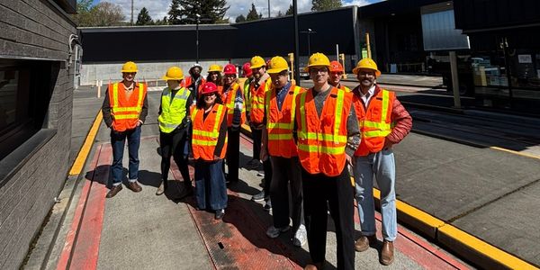 A group of people wearing safety vests and hard hats pose together on an outdoor vehicle scale at an industrial facility on a sunny day.